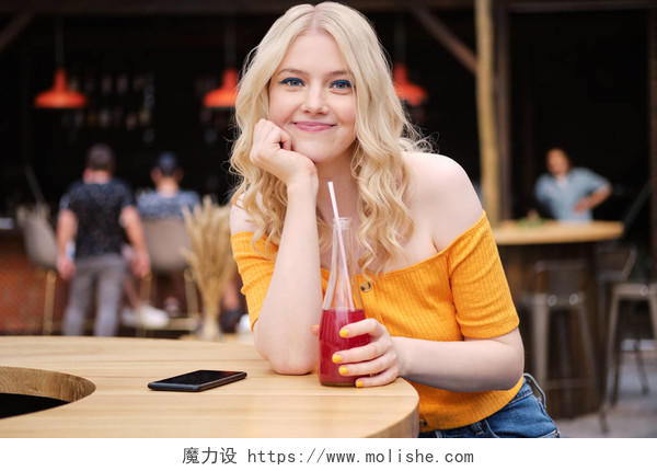 漂亮的微笑金发女孩的肖像愉快地看着相机喝柠檬水在咖啡馆的庭院
